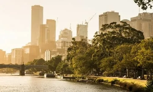 City skyline at sunset along a river, featuring modern skyscrapers and lush trees lining the bank, with a bridge in the foreground.