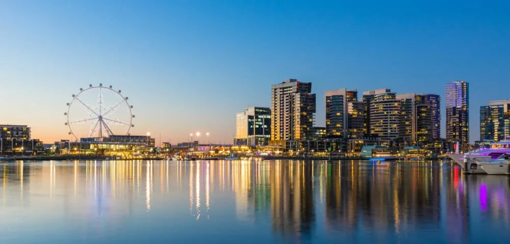 City skyline at dusk featuring a large Ferris wheel and waterfront reflections, showcasing modern buildings and vibrant lights in a lively urban setting.
