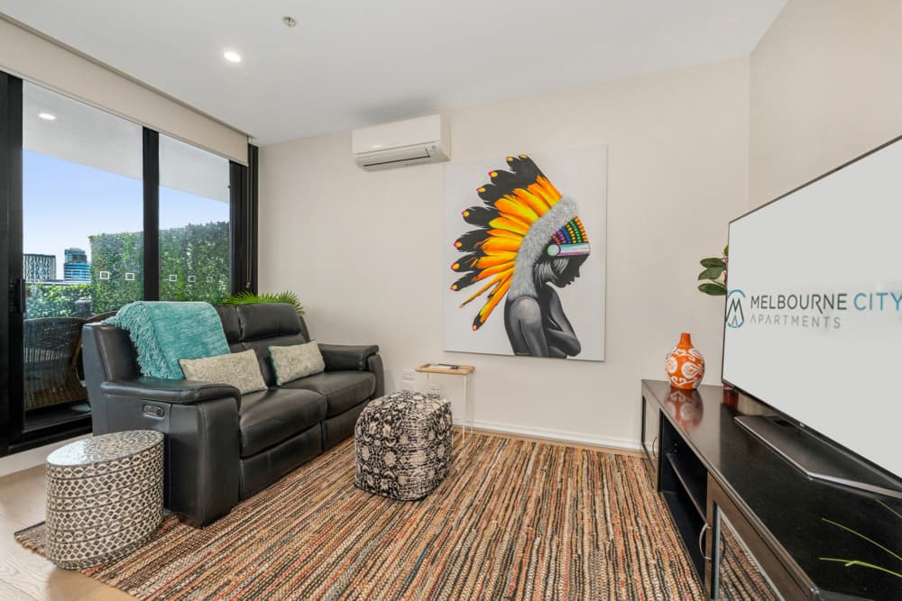 Modern living room featuring a black leather sofa with decorative pillows, a vibrant indigenous-themed artwork on the wall, a stylish pouf, and a decorative vase. Large windows provide natural light and a view of greenery. Ideal for showcasing interior design in Melbourne city apartments.